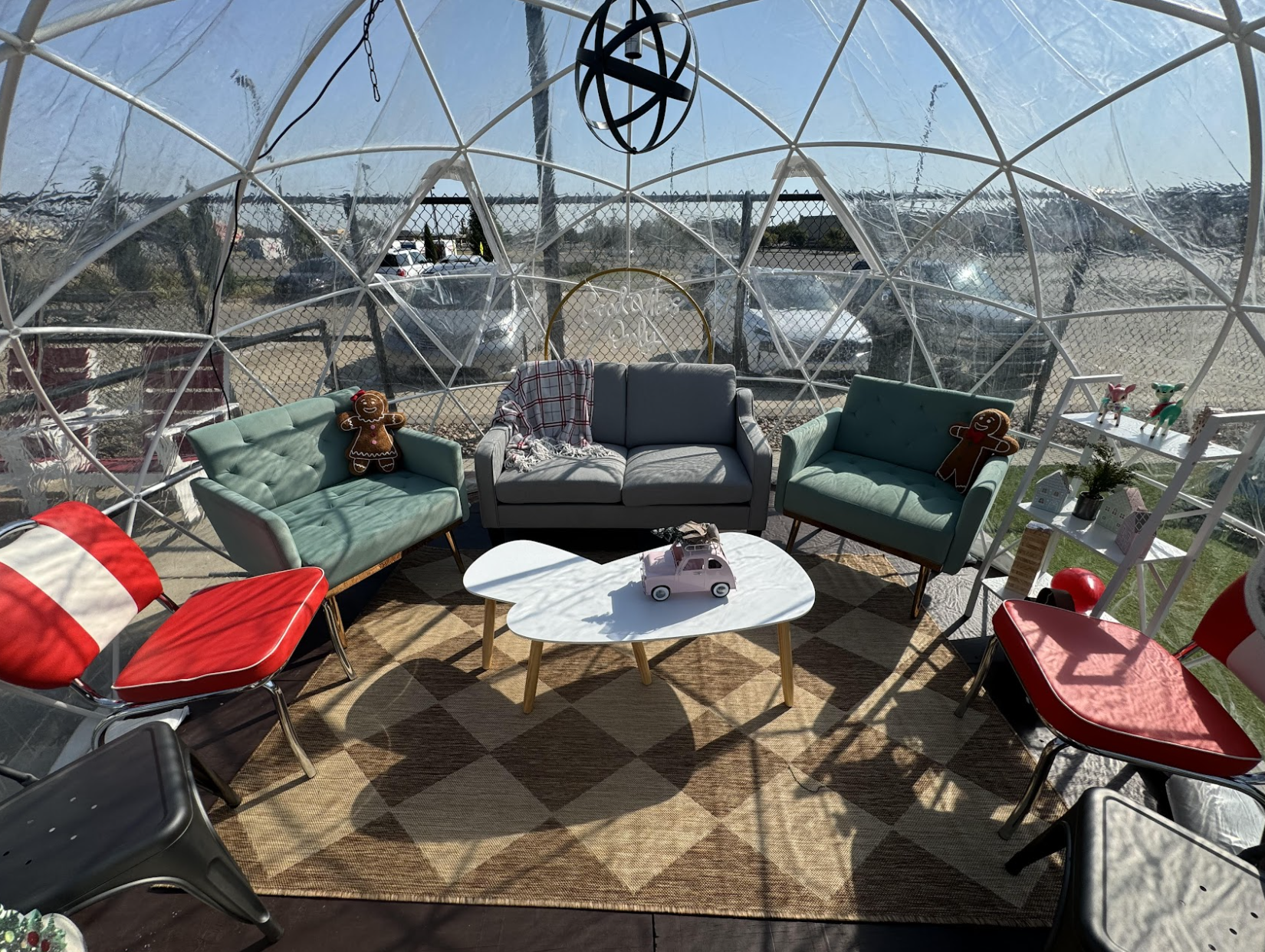 A cozy seating area inside a clear dome with two armchairs, a sofa, red chairs, a white coffee table with toy cars, and gingerbread man plush toys. Sunlight streams in, illuminating the retro decor.